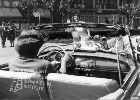 Vlasta Fialová (dr. Emília Kortanová) zdroj: skcinema.sk 
Fotografia z filmu Dom na rázcestí (1959). Za volantom auta sedí Vlasta Fialová (MUDr. Emília Kortanová), v pozadí cez cestu práve prechádza zdravotná sestra s malými deťmi, vzadu možno vidieť budovu s nápisom: KÚPEĽNÁ NEMOCNICA