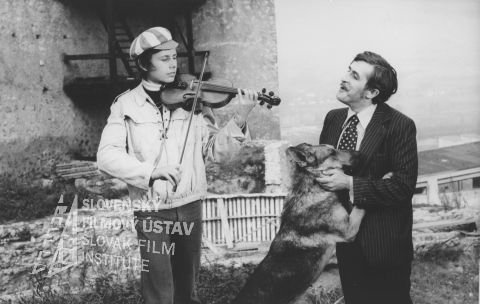 Július Satinský (Pucinko st.) zdroj: skcinema.sk 
Vľavo stojí a hrá na husle chlapec, vpravo stojí so psom Július Satinský (Pucinko st.)